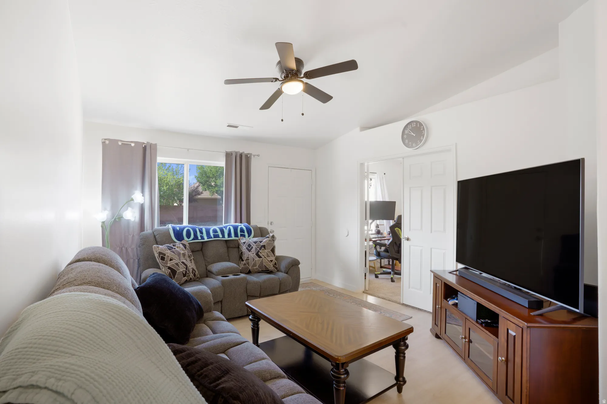 Vaulted ceiling, light wood-finish flooring, ceiling fan with integrated lighting, window with light-filtering drapes, and a white paneled door