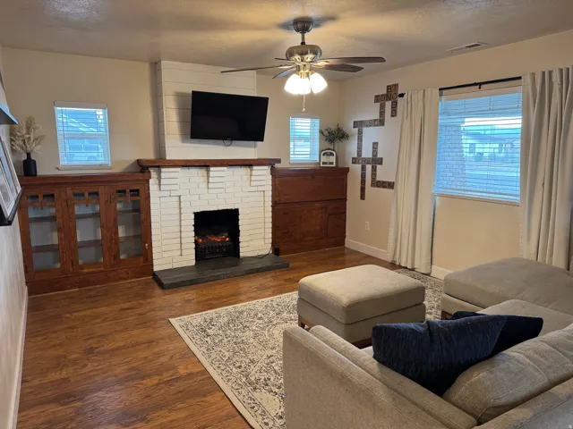 Wood-finish flooring, white brick fireplace with dark stone hearth, built-in wood cabinetry with glass doors, ceiling fan with integrated lighting, and multiple windows with blinds