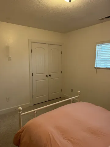 White bi-fold closet doors, wall-mounted light fixture, window with blinds, and neutral carpet flooring