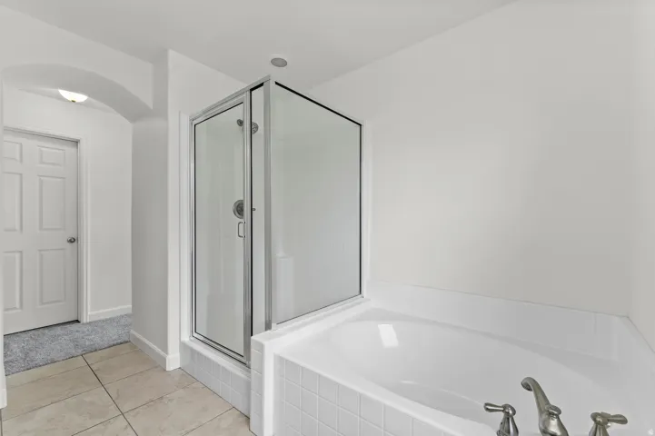 White soaking tub with surrounding tile, separate enclosed shower with clear glass, and neutral-toned floor tiling