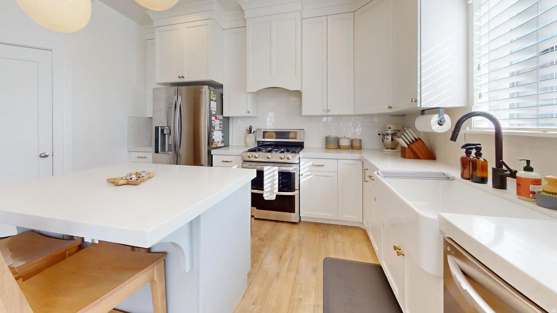 Kitchen featuring white shaker cabinetry, stainless steel appliances, light wood-finish flooring, a white farmhouse sink with a matte black faucet, and an island with a white countertop