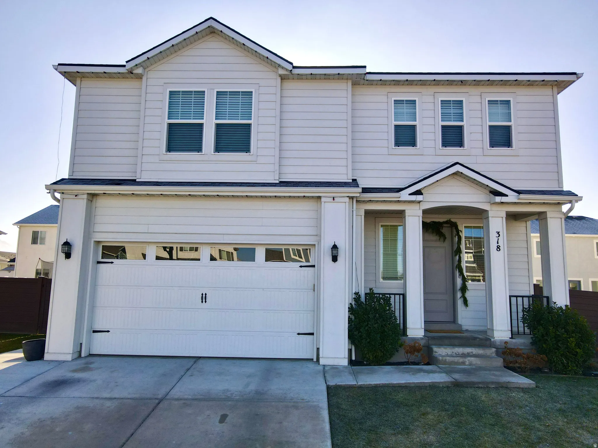 Two-story residence featuring light-colored siding, an integrated two-car garage with transom windows, a covered front porch with columns, a black metal railing, and a gabled roofline