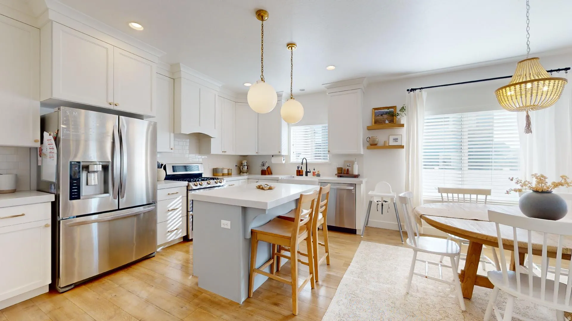 Bright, open-concept kitchen and dining area featuring stainless steel appliances, white cabinetry, a gray center island with wood-finish seating, wood-finish flooring, and pendant lighting