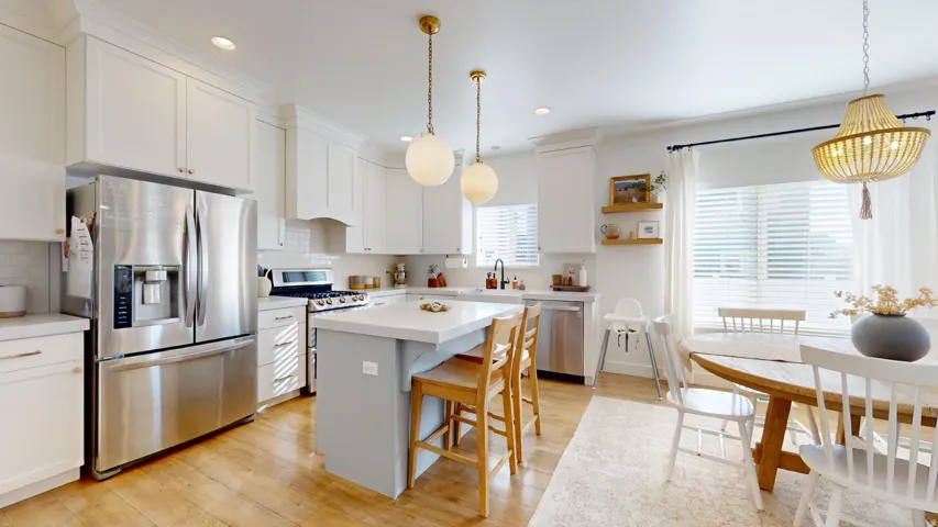 Bright, open-concept kitchen and dining area featuring stainless steel appliances, white cabinetry, a gray center island with wood-finish seating, wood-finish flooring, and pendant lighting