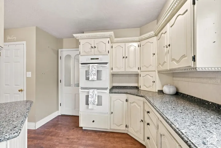 Built-in double ovens, extensive cabinetry with decorative molding, dark speckled countertops, tile backsplash, and wood-finish flooring