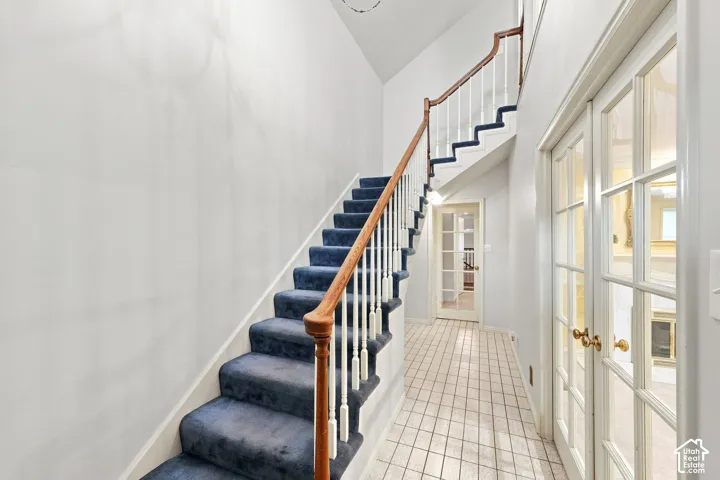Staircase with carpeted treads and wood handrail, white balusters, tiled flooring, and French doors with brass hardware