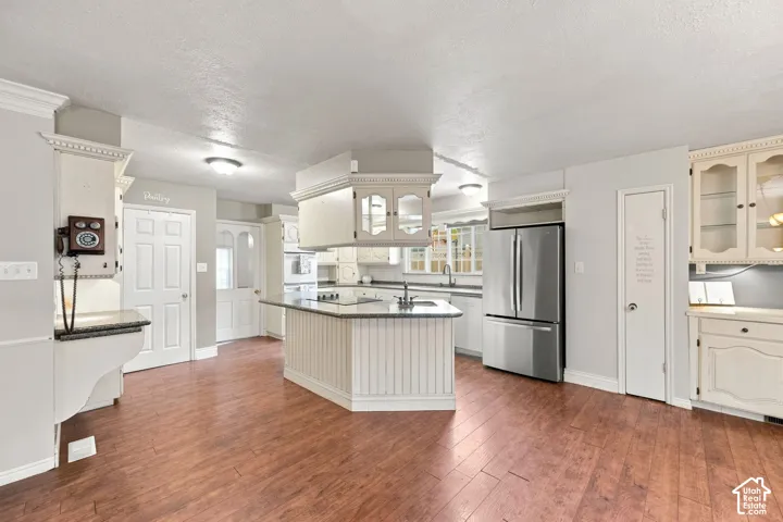 Kitchen featuring wood-finish flooring, white cabinetry, a stainless steel refrigerator, and an island with a built-in sink