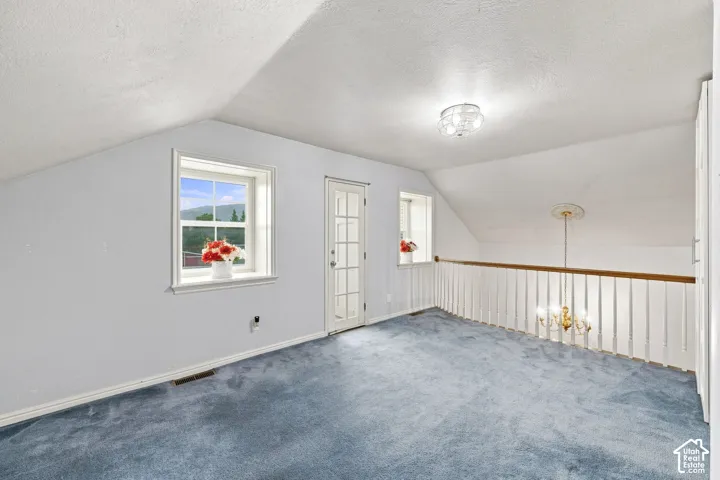 Sloped ceiling room featuring blue carpeting, a white framed window with outdoor scenery, a white paneled door, and a white baluster railing with a wood-finish handrail