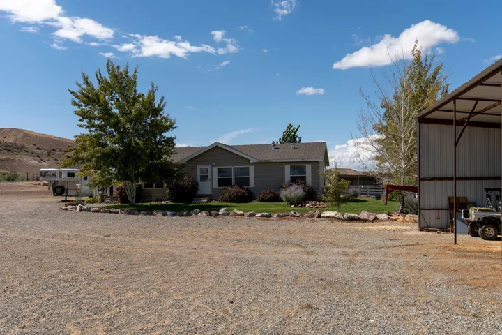 Single-story residence with a pitched roof, gravel driveway, mature trees, and an open-sided metal structure