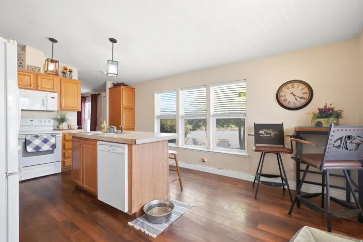 Kitchen featuring wood-finish flooring, a central island with integrated sink, white appliances, and natural wood cabinetry