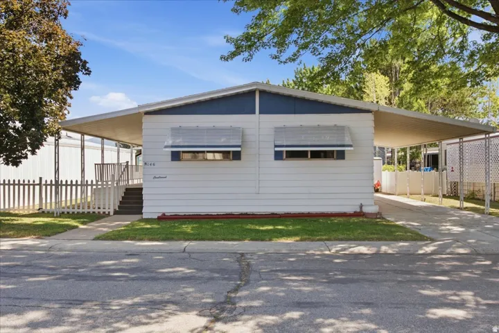 White siding exterior with a contrasting blue fascia, covered entry porch with stairs, white picket fence, and a covered carport with an asphalt driveway