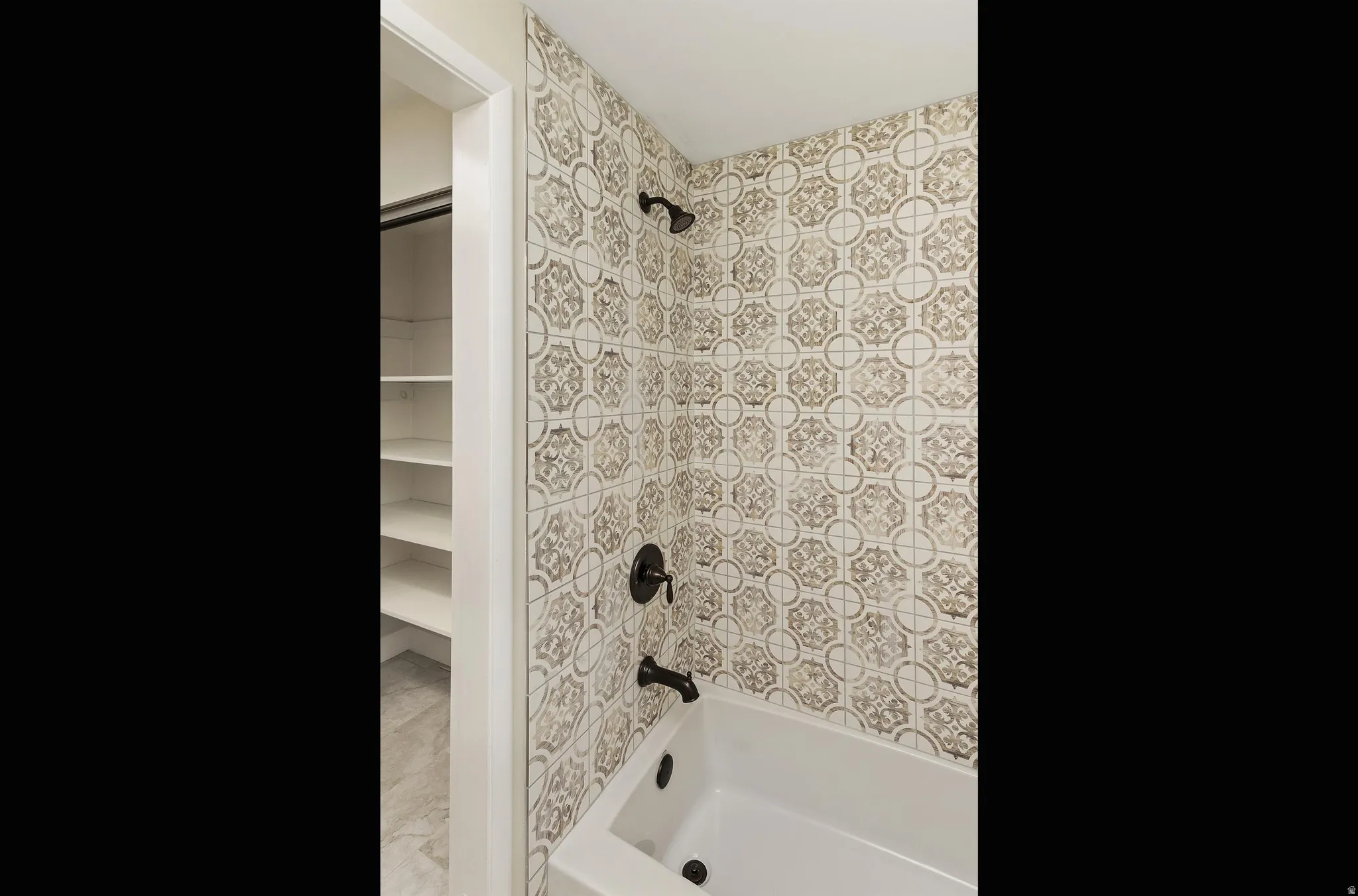 Bathtub with patterned wall tiling, matte black fixtures, and built-in shelving