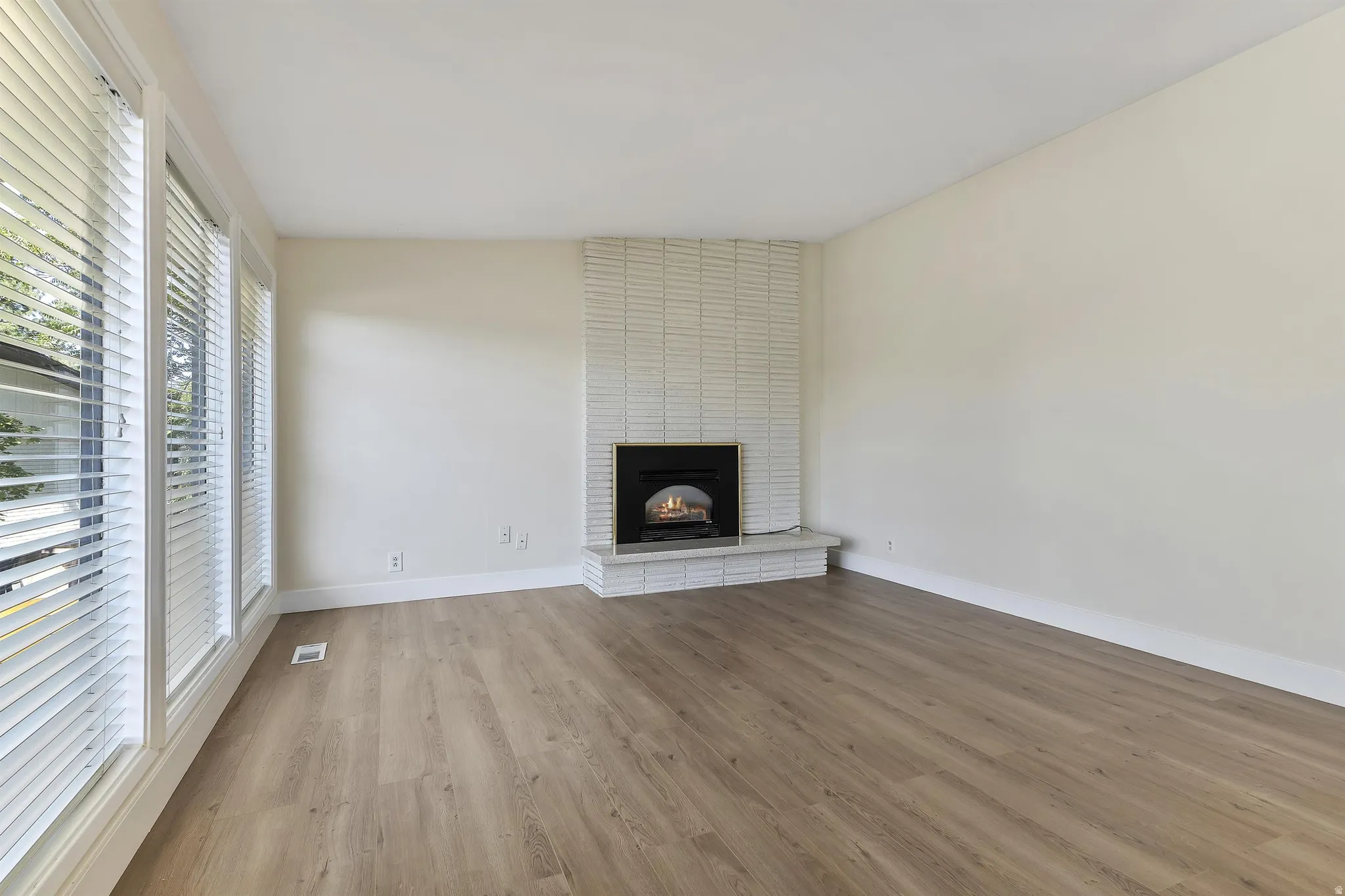 Expansive room featuring wood-finish flooring, a white brick fireplace with a black insert, and multiple windows with horizontal blinds
