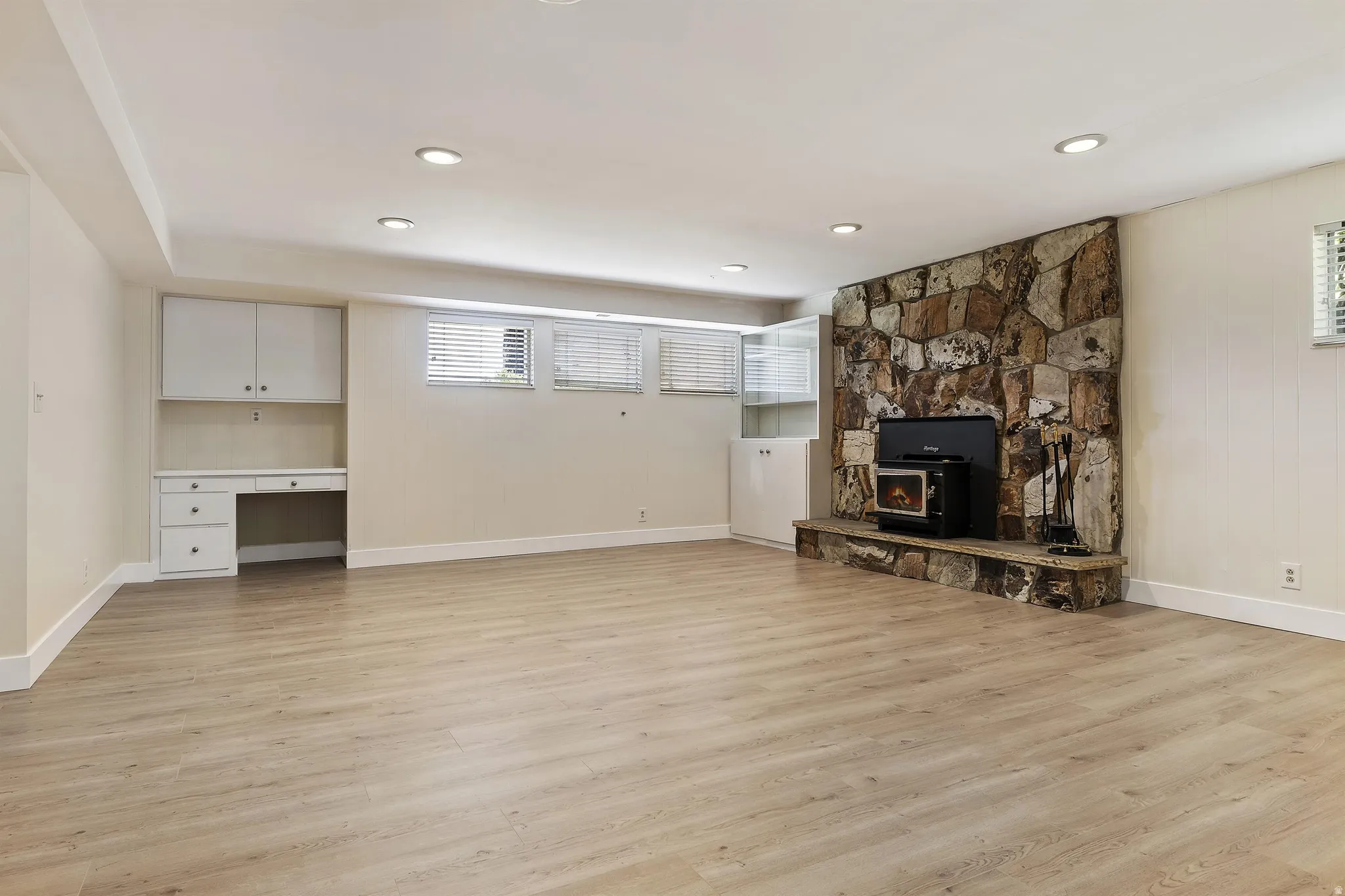 Spacious room featuring wood-finish flooring, recessed lighting, a stone-clad accent wall with a hearth, a wood-burning stove, and built-in desk cabinetry