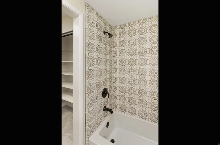 Bathtub with patterned wall tiling, matte black fixtures, and built-in shelving