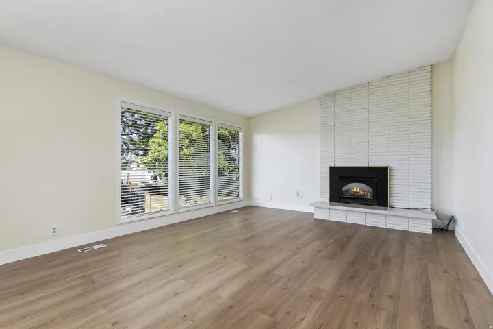 Spacious room featuring wood-finish flooring, a white brick fireplace with a raised hearth, and three large windows equipped with blinds