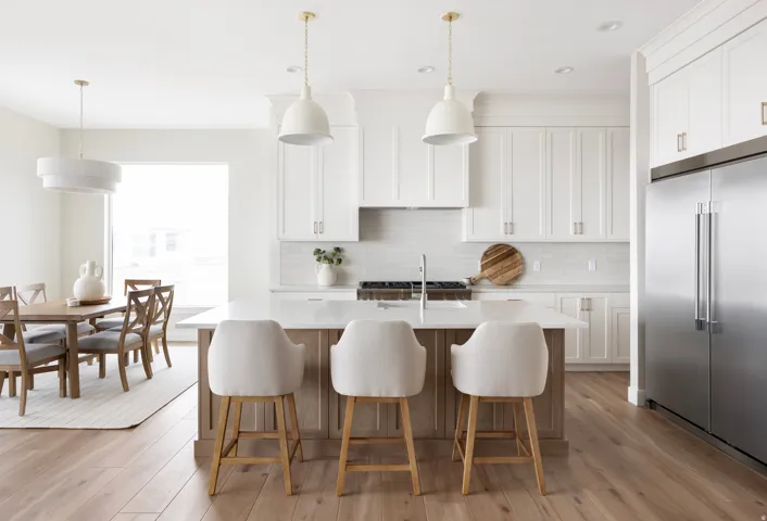 Open-concept kitchen and dining area featuring light wood-finish flooring, white cabinetry with gold-tone hardware, a center island with countertop seating, stainless steel appliances, and pendant lighting
