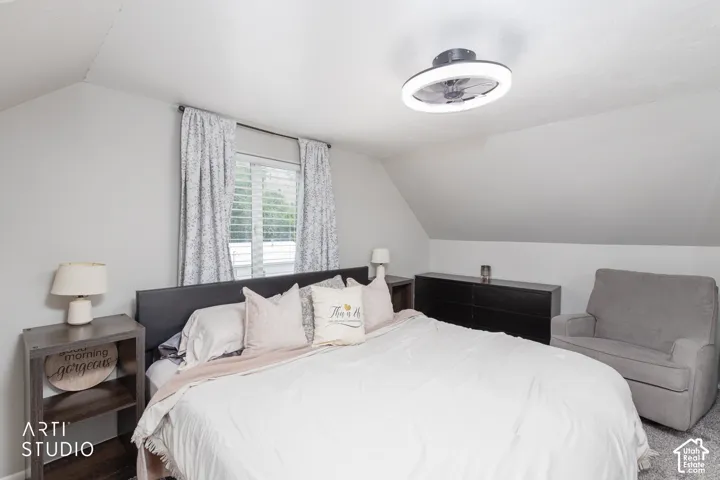Sloped ceiling architecture with a window featuring louvered shutters and drapes, a flush-mount ceiling light with a fan, and wall-to-wall carpeting