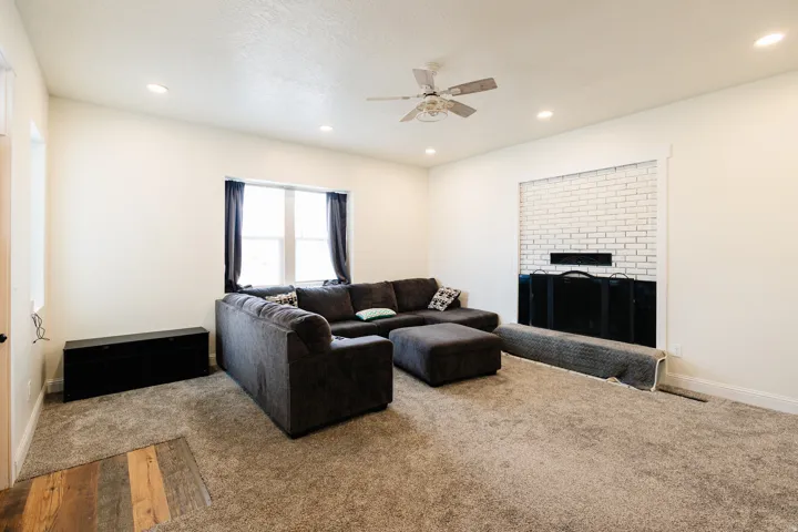 Recessed lighting, ceiling fan, white brick fireplace with black insert, light-toned carpeting, and a window with dark drapery