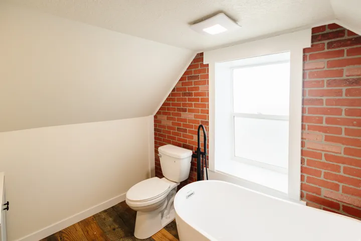 Freestanding bathtub with matte black floor-mounted faucet, red brick wall accents, wood-finish flooring, and a square ceiling light fixture