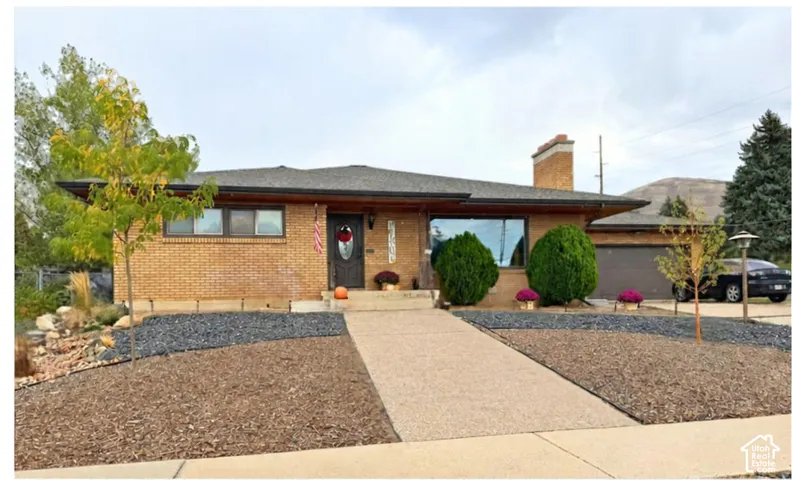 Brick exterior with an attached garage, paved driveway, mature landscaping, and a pitched roof