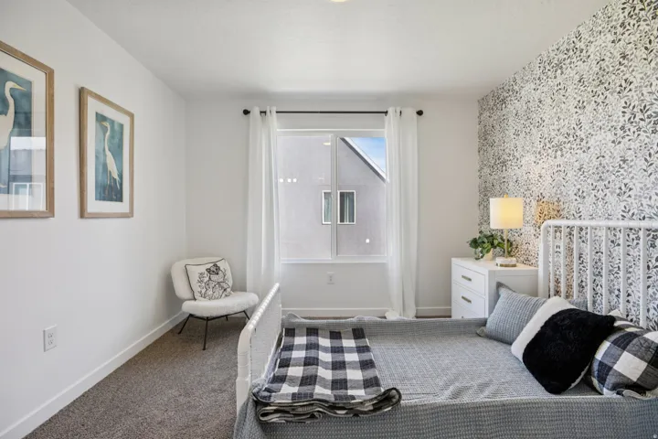 Carpeted room featuring a window with white drapes, a white metal bed frame, a patterned accent wall, and a white nightstand with drawer pulls