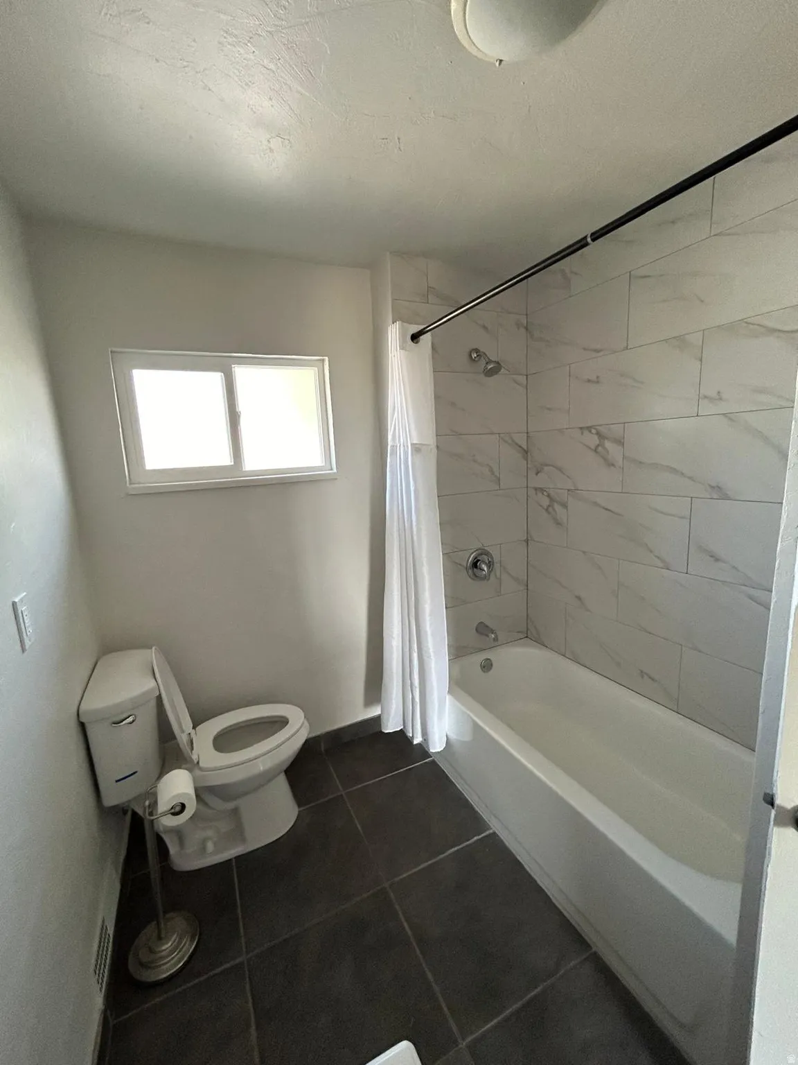 Bathroom featuring a full bathtub with tile surround, a single window, and dark tile flooring