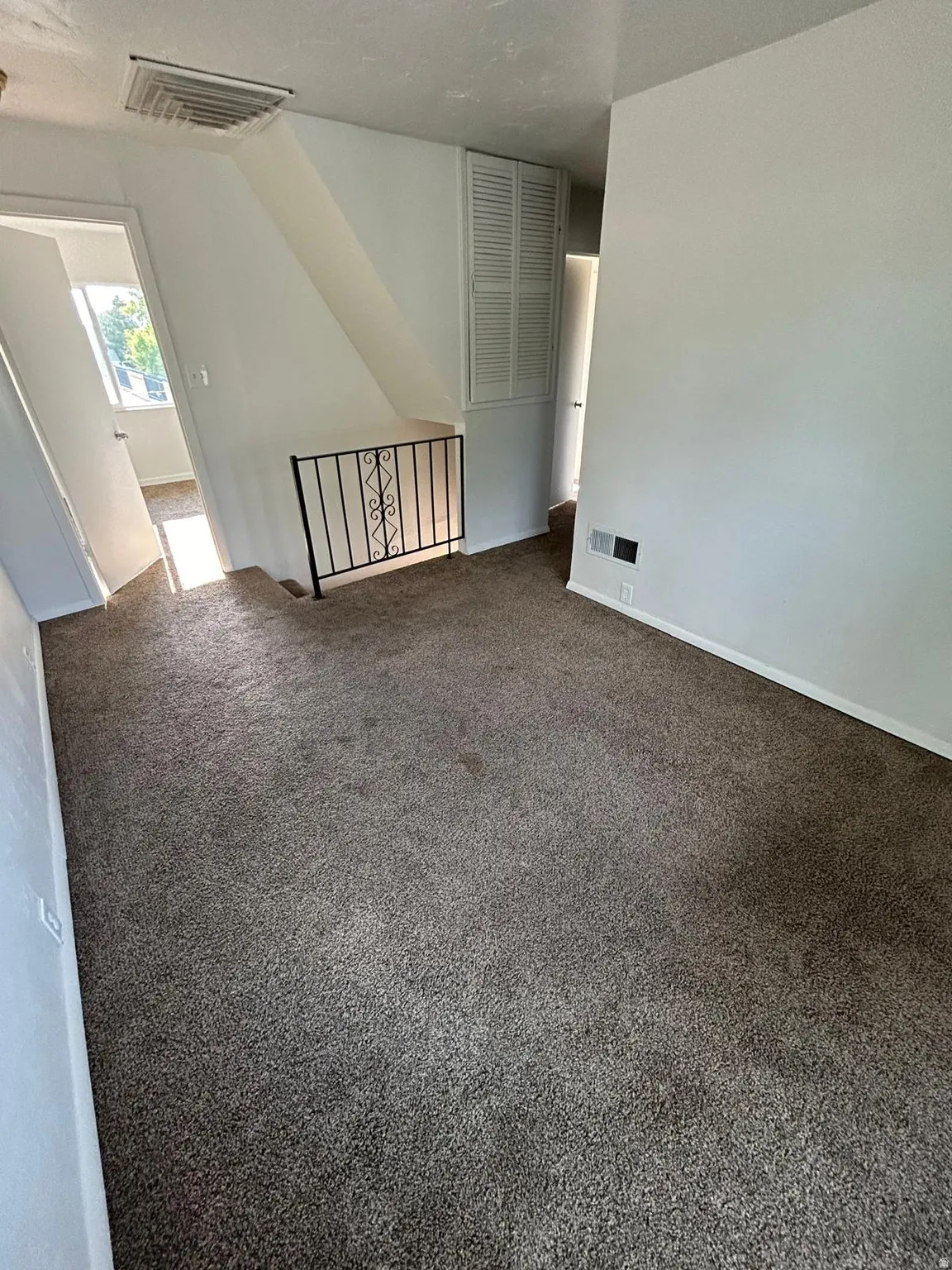 Brown carpeted flooring with white wall finishes, a white louvered door, a black metal railing, and a white ceiling vent