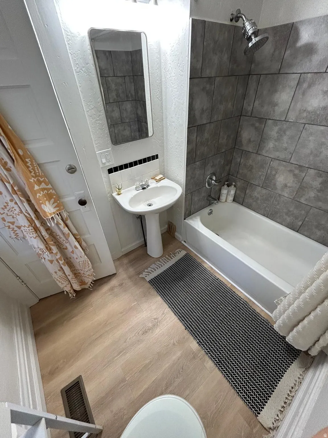 Pedestal sink with chrome fixtures, tiled shower surround, bathtub with chrome showerhead, wood-finish flooring, and wall-mounted mirror