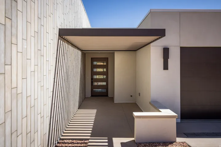 Modern exterior featuring a prominent wood entry door with five glass panels, a dark metal overhang, and a textured light-colored vertical paneled wall