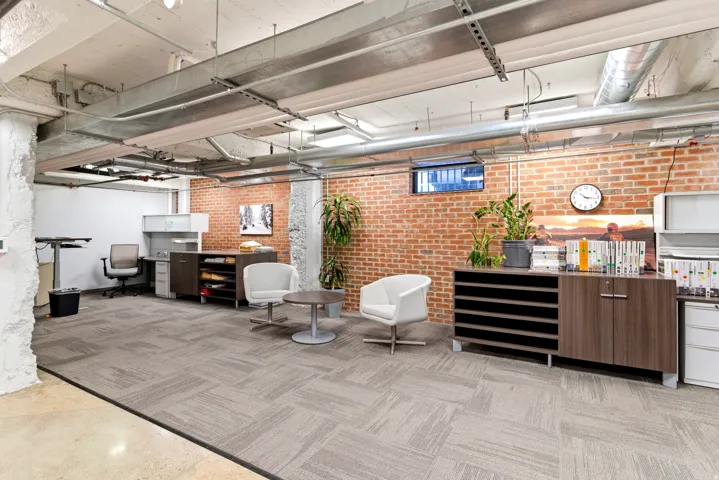 Exposed brick wall, industrial-style ceiling with visible ductwork, patterned carpet tile flooring, and a built-in wood-finish storage unit