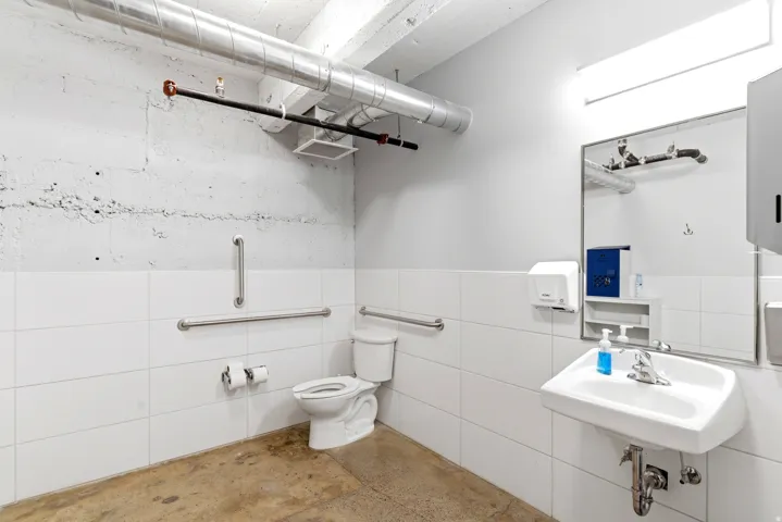 Restroom featuring a wall-mounted sink, toilet, white tile wainscoting, exposed ceiling ductwork, and grab bars
