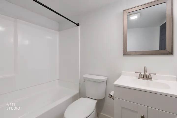 White tub shower combination with black shower rod, white vanity with integrated sink, brushed nickel faucet, framed mirror, and white toilet