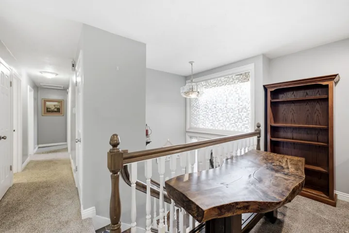 Upper-level landing featuring elegant wood-finish railing with turned balusters, solid wood-finish table with natural edge detailing, built-in wood cabinetry with glass-front shelving, recessed and pendant lighting, and carpeted flooring throughout the sp