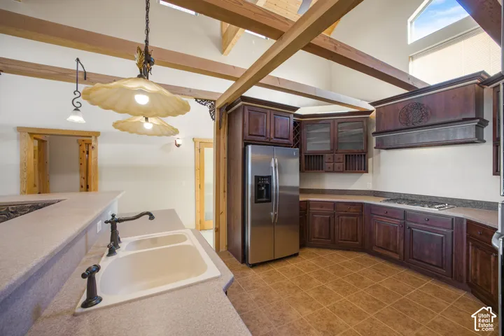 Vaulted ceiling with exposed wood beams, stainless steel French door refrigerator, gas cooktop, tile flooring, and a double basin sink with a bronze-finish faucet