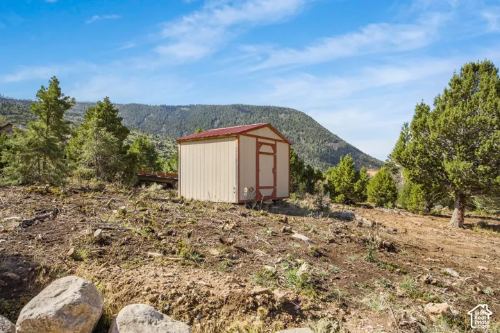 Detached storage shed with a red metal roof and beige siding, set against a backdrop of mature evergreen trees and distant mountains