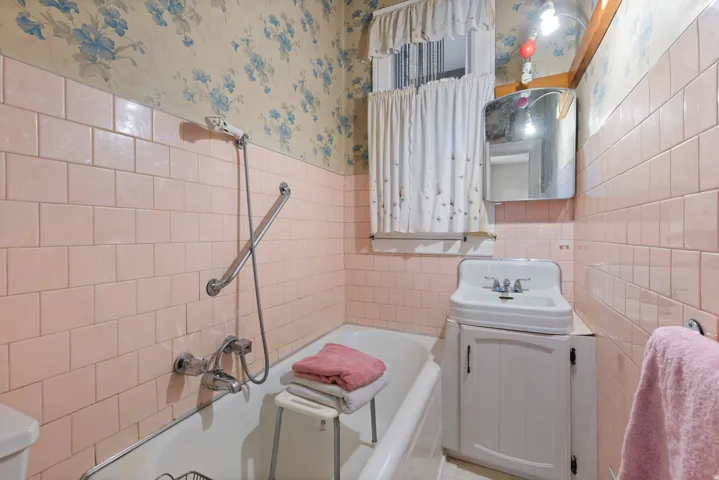 Bathroom featuring a built-in bathtub, wall-mounted shower fixture, ceramic wall tiling, and a pedestal sink with a storage cabinet
