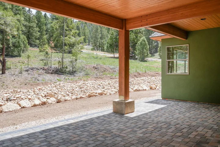 Covered patio with exposed wood beam ceiling and stone-finish flooring, opening to landscaped grounds featuring native evergreen trees, river rock hardscaping, and natural terrain
