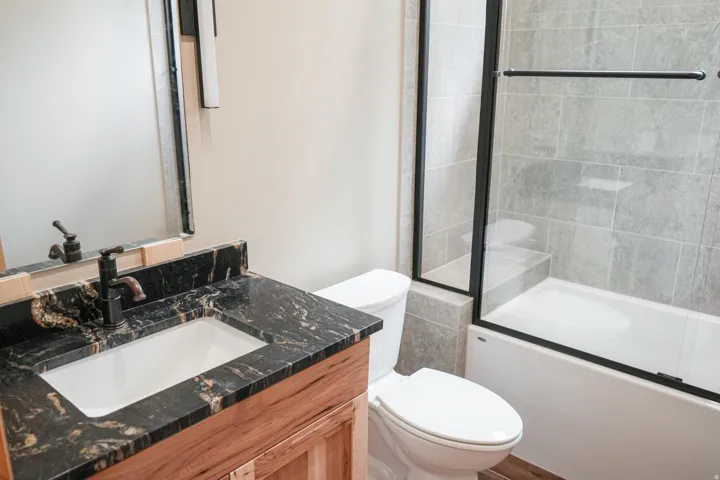 Black granite vanity countertop with integrated sink, wood-finish vanity cabinet, shower/tub combination with sliding glass doors, and light gray tiled shower surround