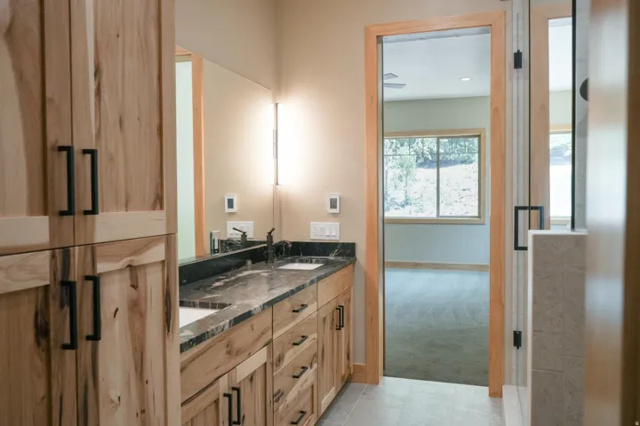 Dual vanity with a dark countertop and undermount sinks, custom wood cabinetry with matte black hardware, and a glass-enclosed shower with tiled walls