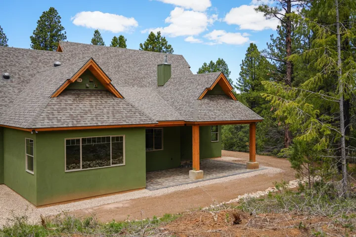 Green stucco exterior with a gray shingle roof, wood-wrapped gables, and a covered patio featuring wood support columns and paver flooring