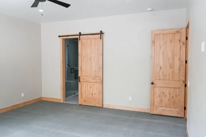 Neutral gray wall paint with light wood trim and matching doors, dark gray patterned carpeting, and a ceiling fan with integrated lighting