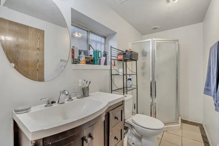 Vanity with an integrated sink, polished chrome faucet, round wall mirror, corner shower with frosted glass, and tile flooring