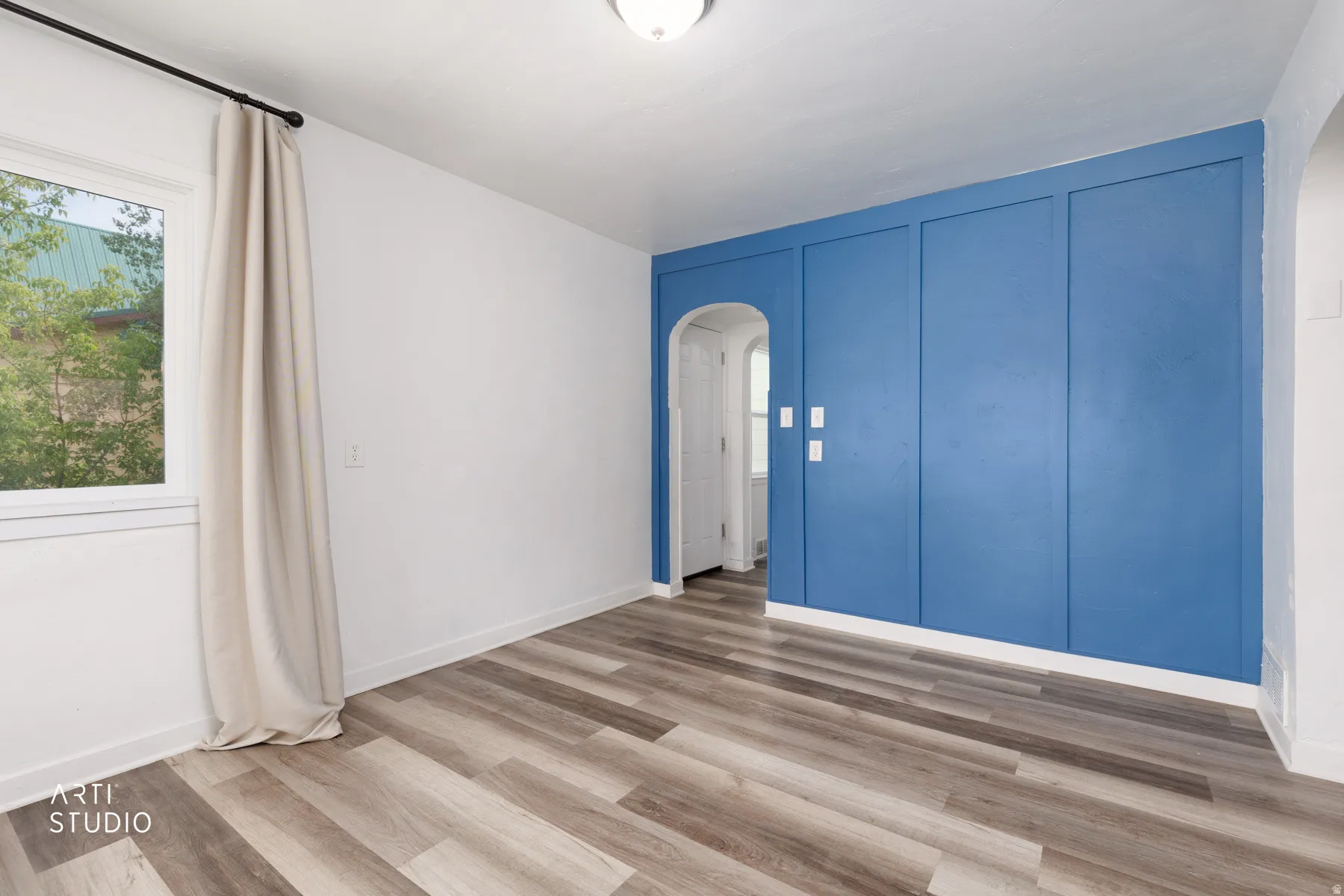 Wood-finish flooring, accent wall with paneling, white window trim, arched doorway, and overhead ceiling light fixture