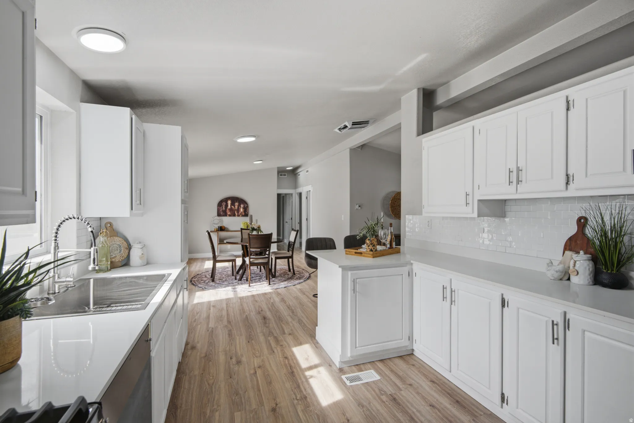 White cabinetry with brushed nickel hardware, stainless steel sink, subway tile backsplash, light-toned countertops, and wood-finish flooring