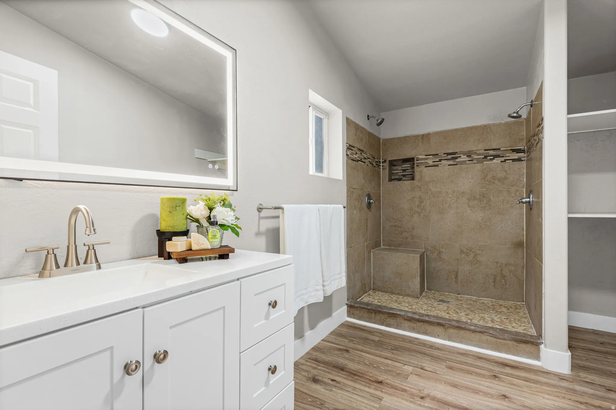 Vanity with integrated sink, illuminated mirror, wood-finish flooring, tiled shower with built-in bench, and shelving