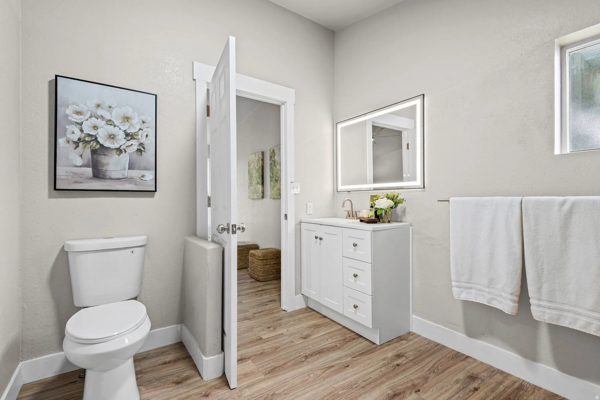 Wood-finish flooring, white vanity with cabinet and drawer storage, illuminated mirror, and a modern toilet
