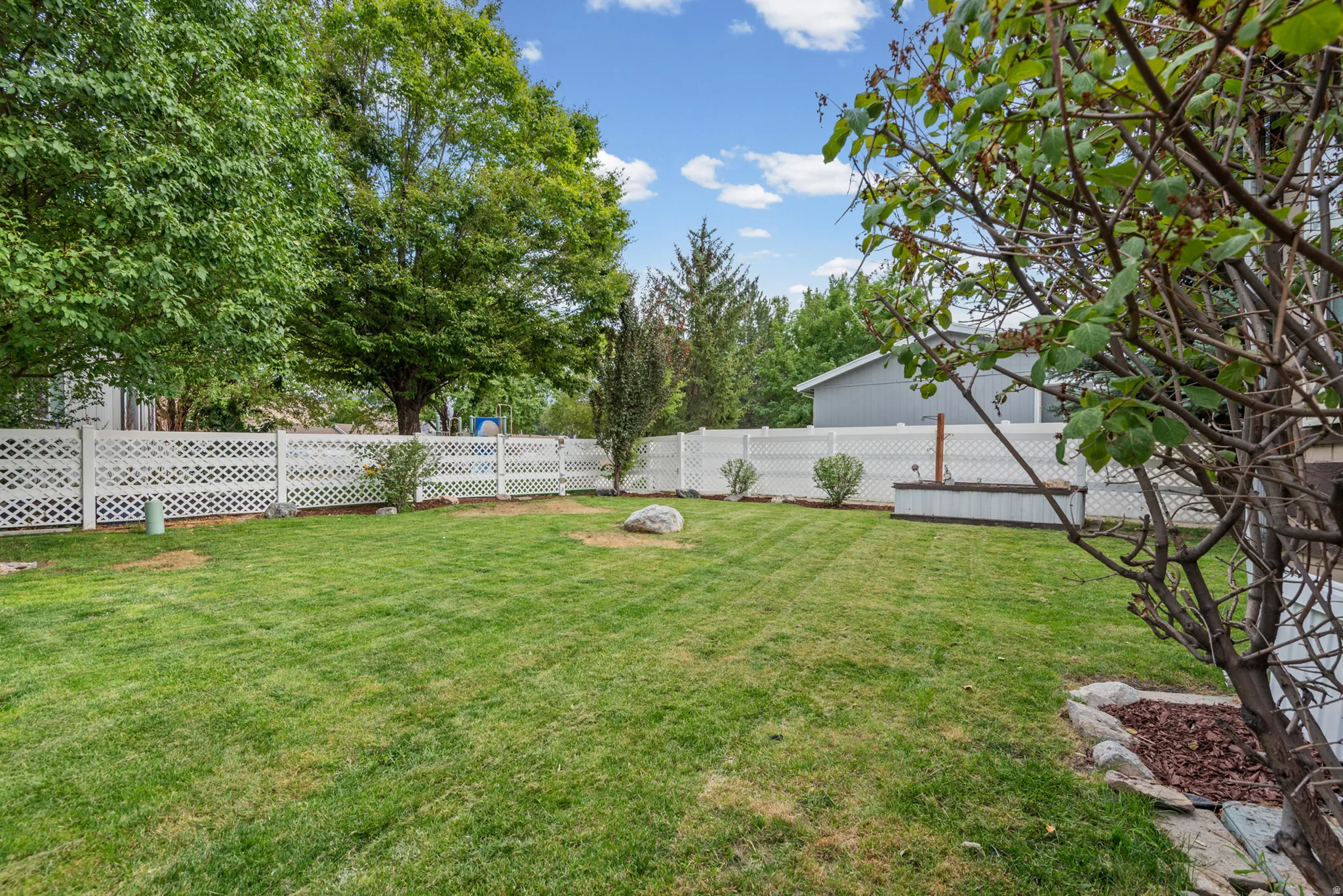 Expansive backyard featuring a green lawn, white lattice fencing, mature trees, and a raised garden bed