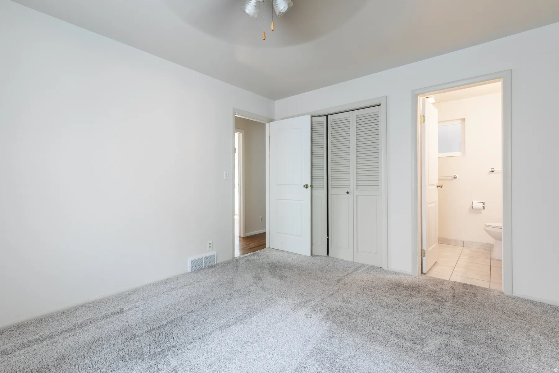 Plush carpeted flooring, a ceiling fan, louvered bi-fold closet doors, and an attached bathroom with tiled flooring