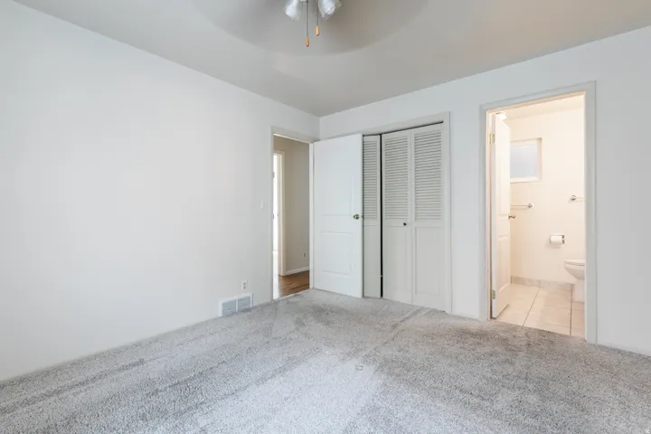 Plush carpeted flooring, a ceiling fan, louvered bi-fold closet doors, and an attached bathroom with tiled flooring
